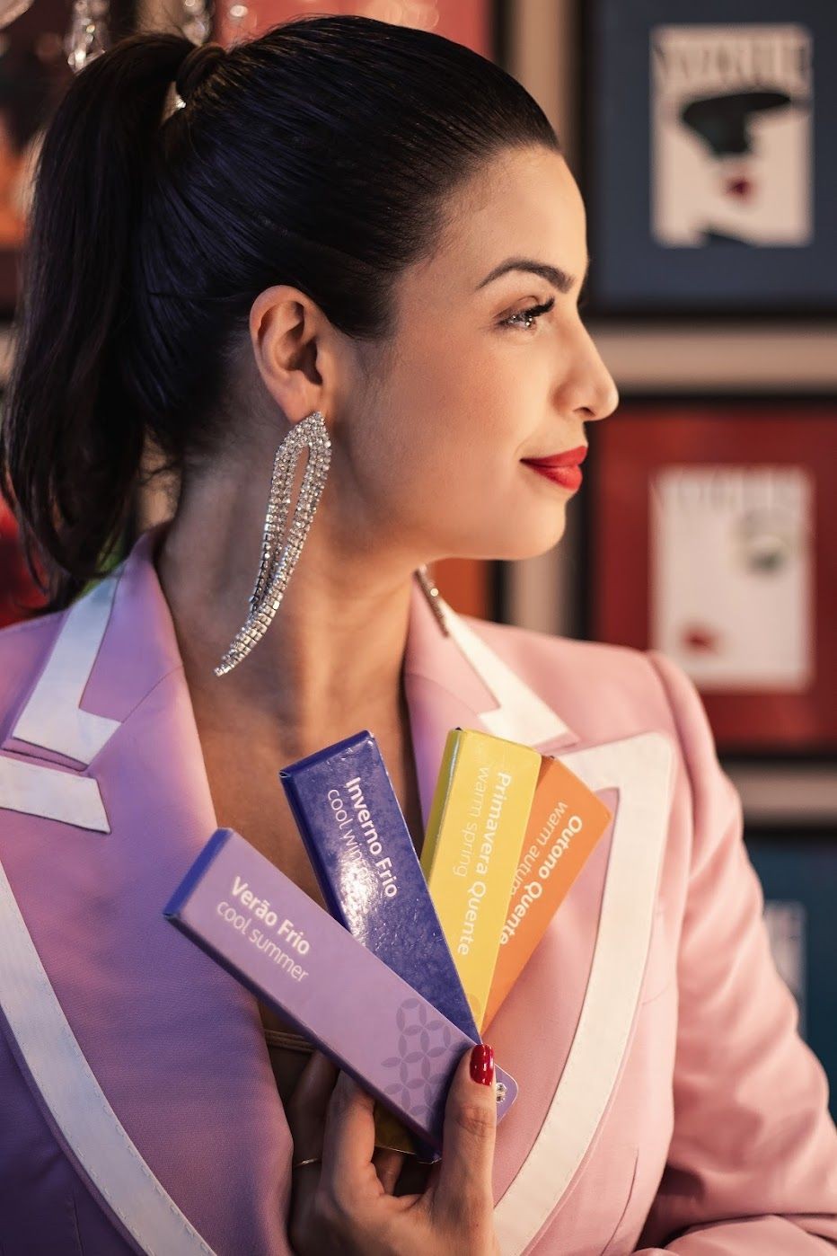 Close-up photo of an elegant woman in her thirties, wearing earrings and a pink blazer with white details, holding colorful business cards inside a room. The color scheme is symbolic, with a palette of pink, purple, and orange, and red walls and books. The jewelry is elegant, and the photography style is editorial, reminiscent of a Vogue magazine cover, with flash lighting, volumetric light, daylight, and volumetric shadows