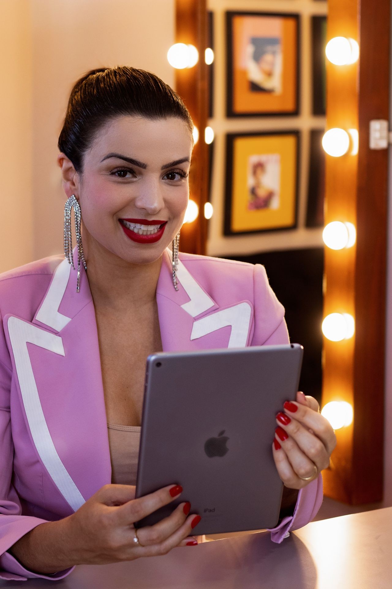 A candid photo of an elegant woman in her late thirties, wearing bold pink and white blazer with shoulder pads holding up the new Apple ipad to camera, red lipstick, diamond earrings, sitting behind desk on stage at talk show studio set, smiling, yellow lighting, black background, luxury aesthetic , real skin details